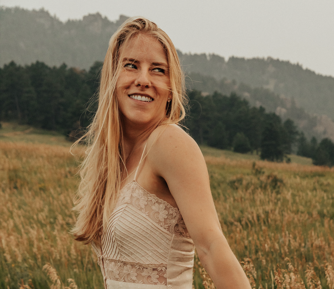 Woman with blonde hair standing in a field with mountains in the background